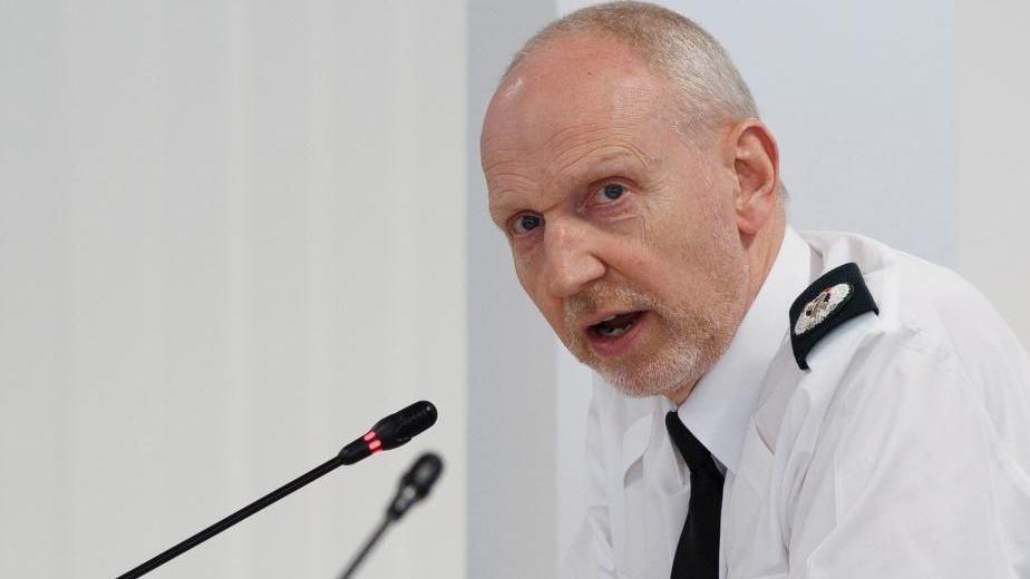 Davy Beck speaking into a microphone at a policing board meeting. He's wearing a white shirt and black tie.