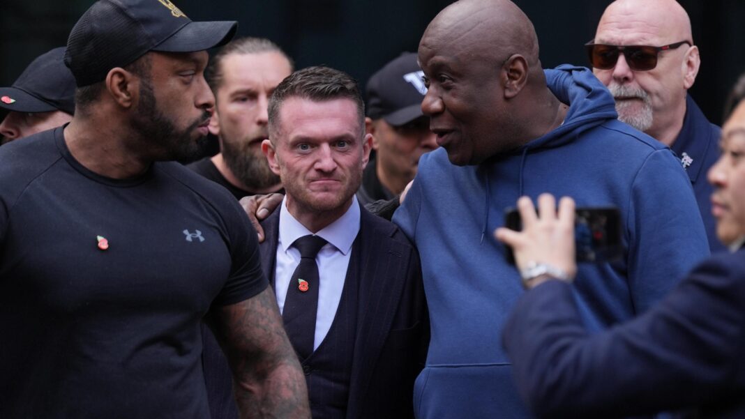 Robinson is wearing a suit and a poppy badge on his tie, he is surrounded by a large group of men. One tall man has his arm around Tommy as he speaks to him