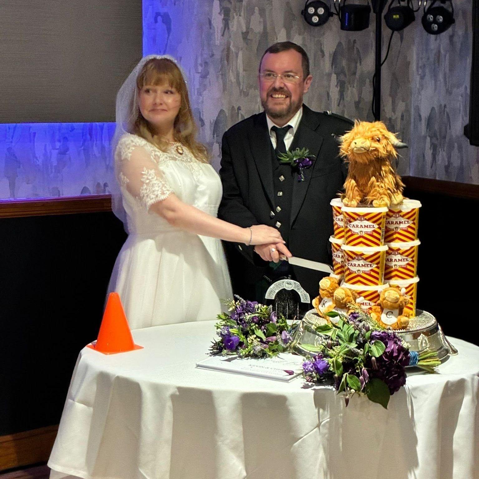 A bride and groom cut their wedding cake which sits on a table and is made of tubs of caramel logs, with a highland cow on top.