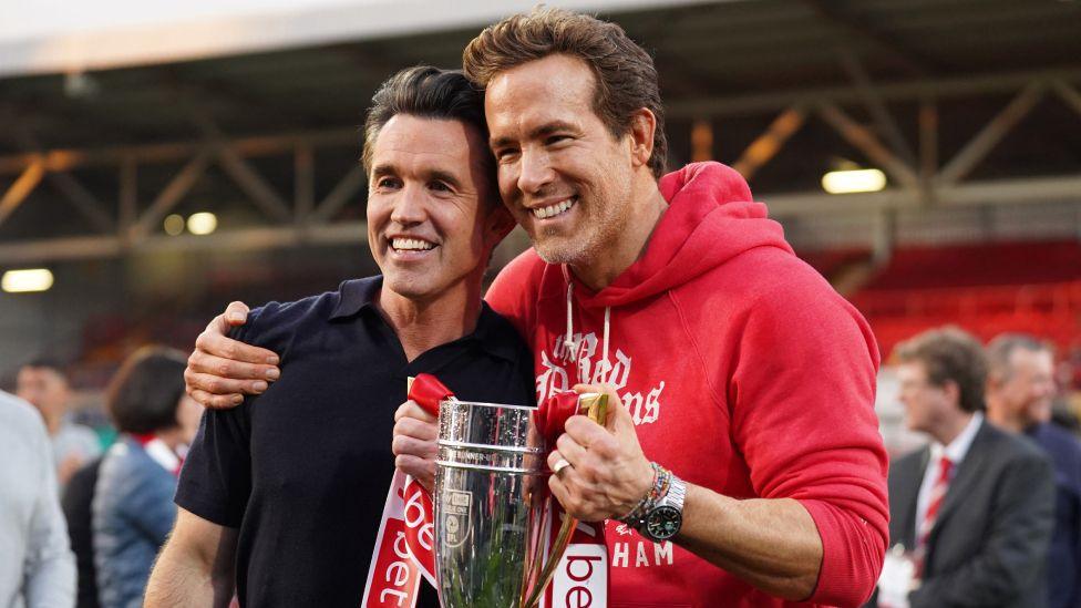 Rob McElhenney and Ryan Reynolds, co-owners of Wrexham AFC, pose for a photo with the trophy following the club's promotion to the Sky Bet Championship during the Sky Bet League One match between Wrexham AFC and Charlton Athletic FC at Racecourse Ground on April 26, 2025 in Wrexham, Wales