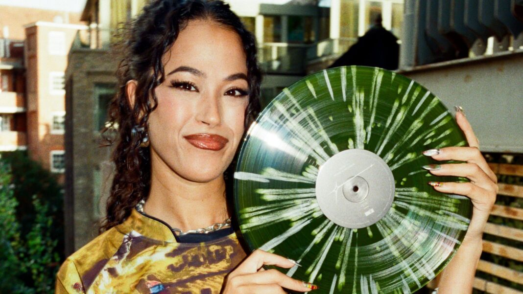 Jazzy pictured with her debut album on a green vinyl. She smiles at the camera holding the LP, her hair in a ponytail. She's wearing a yellow sport-style shirt and is pictured outside with flats visible behind her.