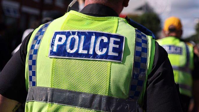 A police officer walks in a crowd alongside other officers. He has an ear piece and is wearing a fluorescent yellow tabard with Police written on the back