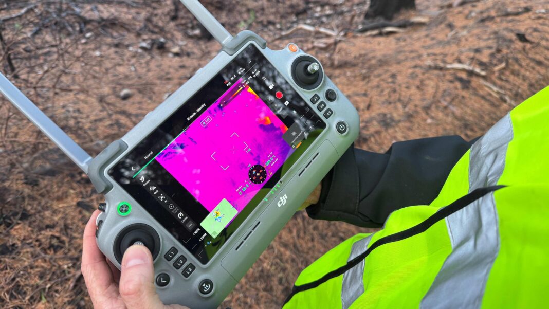 A man's hands holding a drone controller with a thermal image displayed on the screen in bright purple.