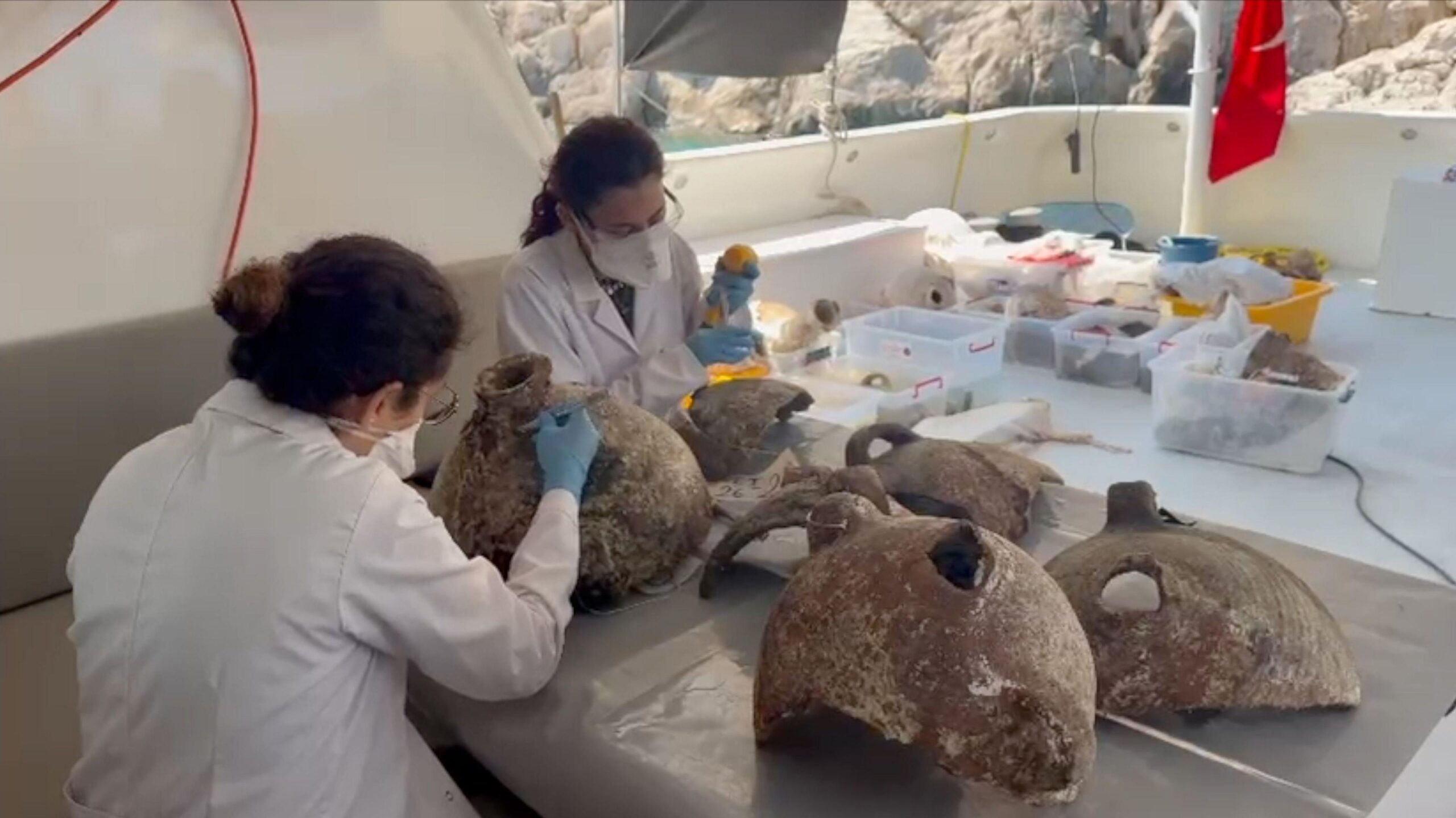 Two people in lab coats and wearing blue gloves and masks are examining some of the amphorae that have been brought to the surface. They are sitting at a table at the back of a dive boat and are in the shade under the boat's roof. The person on the left has her hair tied up and is looking at an intact amphora. She is gently and carefully scraping the surface close to the neck with a scalpel. There are 4 other partial amphorae on the table. Another person who sits slightly behind and to the left, is looking at one of the other pieces of pot. In the background, on the deck of the boat are 6 plastic crates that contain more pieces of pottery brought up from the sea floor. The rocks of the coastline can be seen just behind the stern of the boat and part of a red flag that is attached to a pole on the boat is hanging down in the top right hand corner.