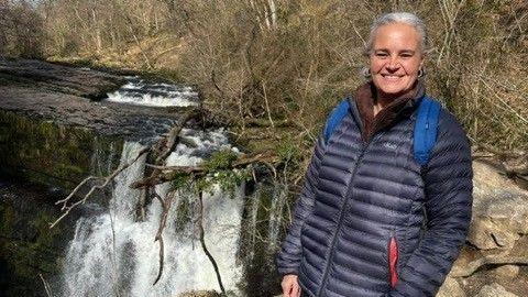 Eleanor stands by a waterfall. She has white hair, which is tied back, and wears a navy coat. She is smiling at the camera.