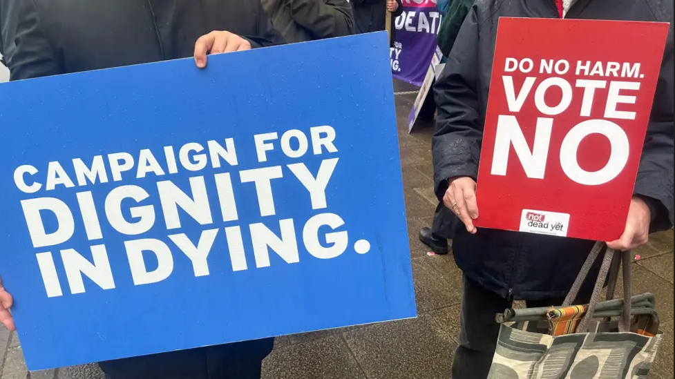 A blue campaign for dignity in dying sign on the left, next to a do no harm, vote no sign on the right.