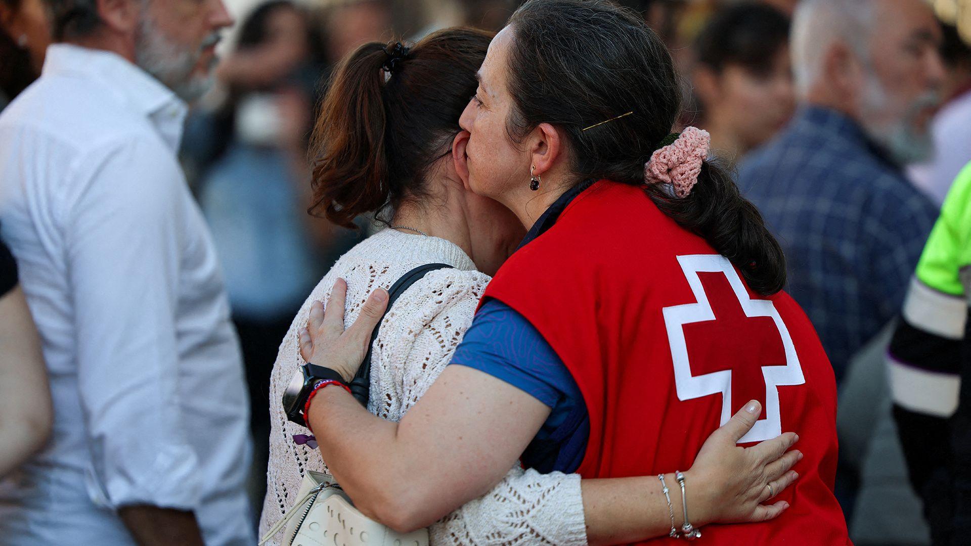 A person in a paramedic vest hugs another person next to a group of people while waiting outside crash scene in Urda on Wednesday