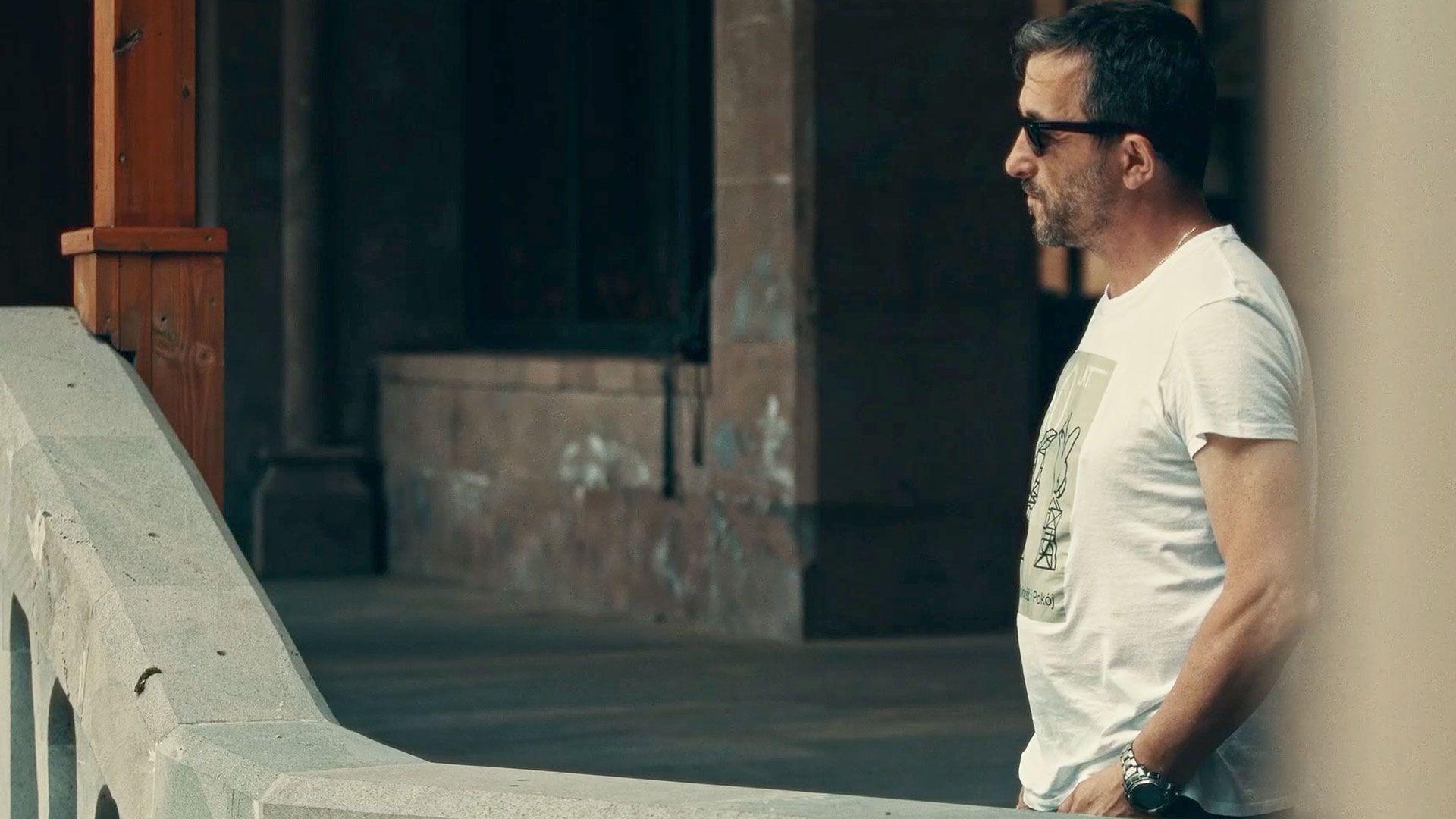 Marcin Mamon is wearing a white t-shirt and dark glasses. He is standing with his hands in his pockets waiting by a stone wall.