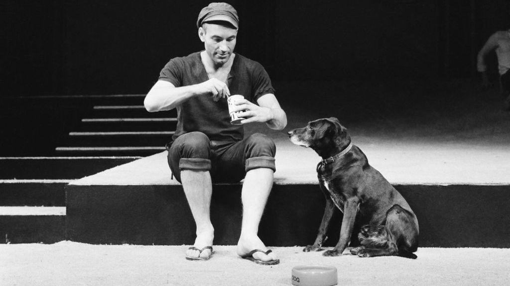 Sir Patrick on stage with a dog that he is feeding as part of a stage play. He is wearing a hat a sandals.