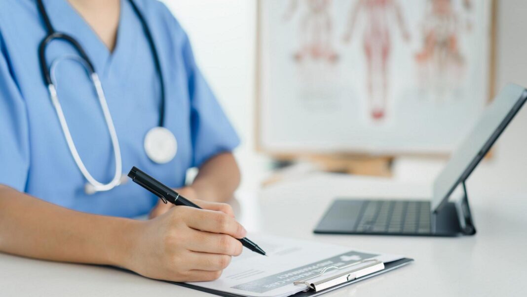 Consultant doctors in England to strike next month A nurse is writing on a clipboard with a black pen. There is a laptop in front of the nurse. On the table is a laptop. The nurse has blue overalls on. The background is blurred but there is a chart on display.