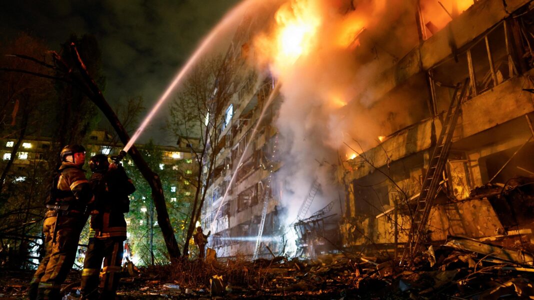 Two firefighters hold a hose spraying water onto an apartment building which is alight