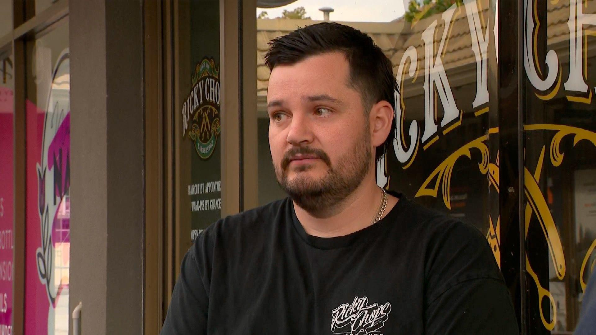 A man dressed in a black T-shirt with a beard and brown hair standing out the front of his barber shop.