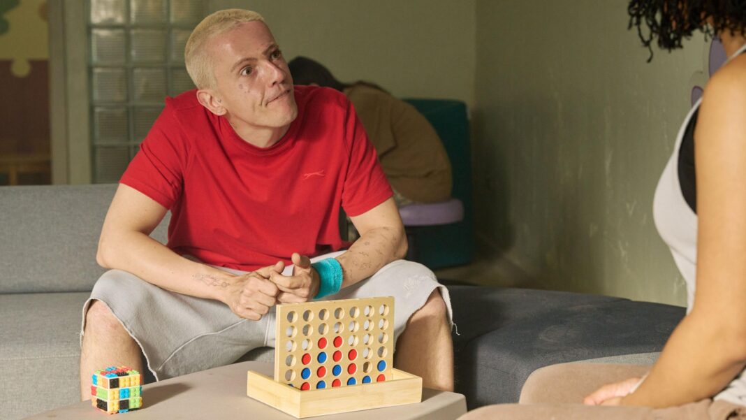 Robert Rhodes, as Sonic, in television show The Rapture has short blonde hair, is wearing a red T-shirt, pale shorts and has a wooden game in front of him. A woman is to the right.