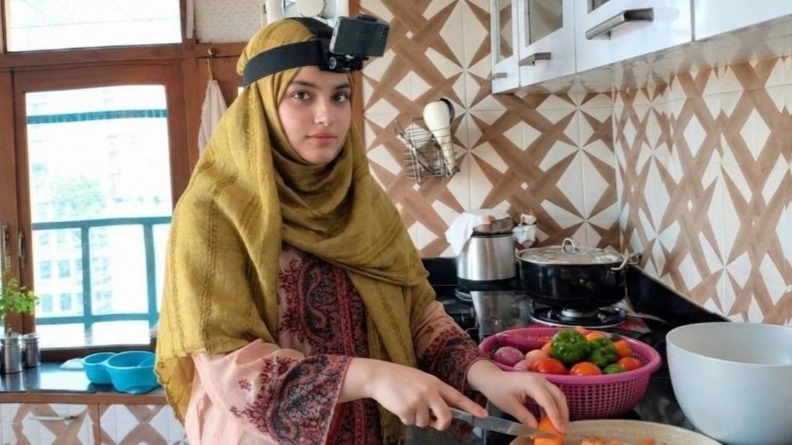 Jolly Arifur wears a mustard-coloured head scarf with a camera attached. She is chopping a carrot in her kitchen.