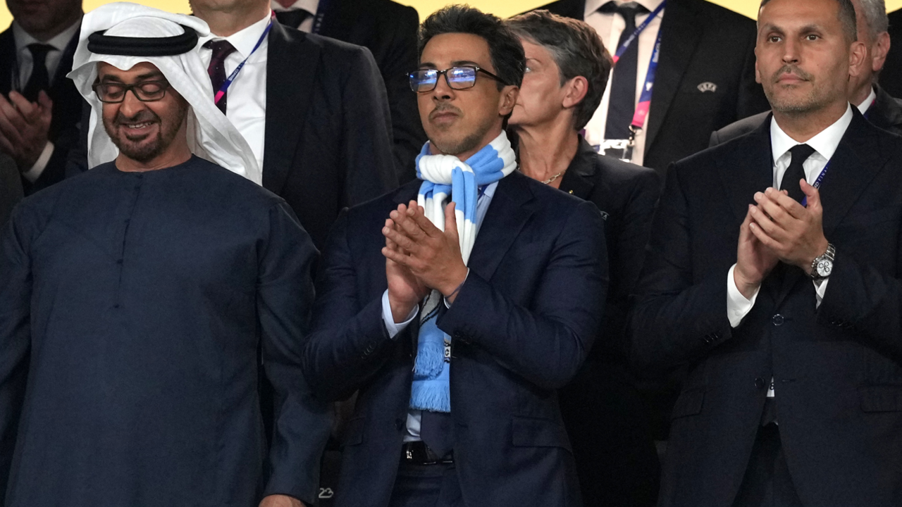 Image shows Sheikh Mansour bin Zayed Al Nahyan, wearing a Man City scarf, and standing in the stands at the Etihad stadium.