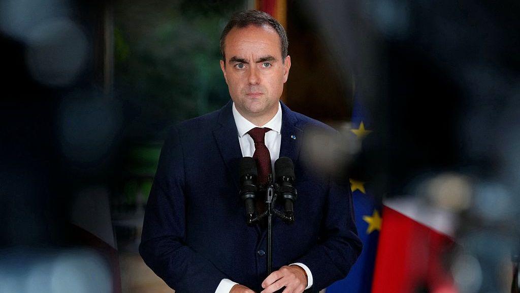 A sombre man in a blue suit and red tie speaks before two microphones in front of a blurred background.