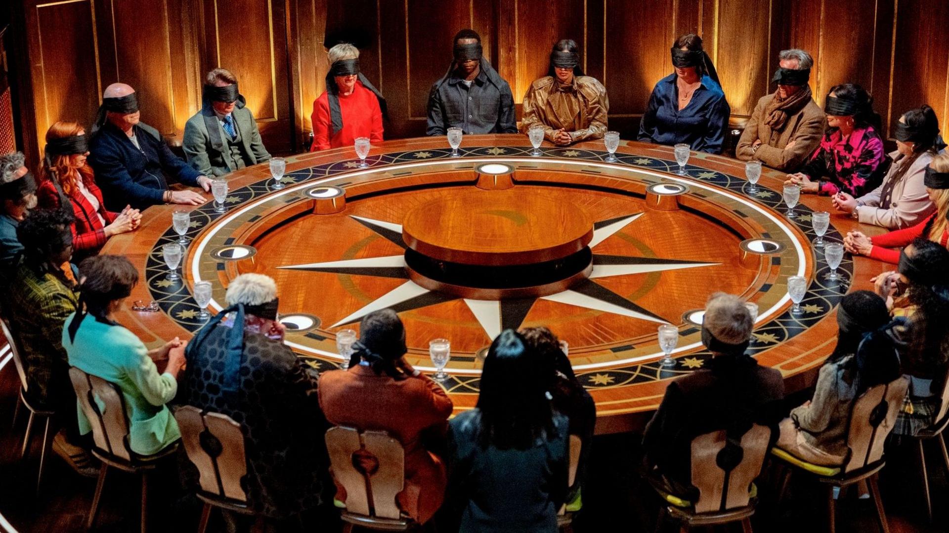 A screen shot from the Celebrity Traitors season 2. A group of 21 people sit around a large round table. They are all wearing blindfolds.