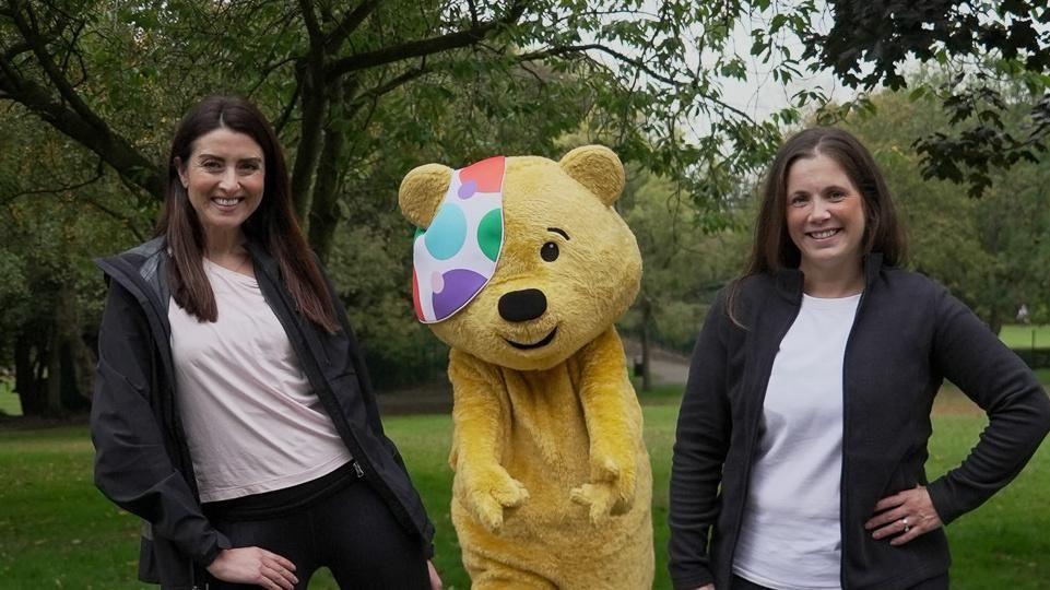Two adults stand on either side of a person in a yellow bear costume with a colourful patch over one eye. The group is outdoors in a grassy, tree-lined park. All three are smiling at the camera, and the adults are dressed casually in jackets and shirts.