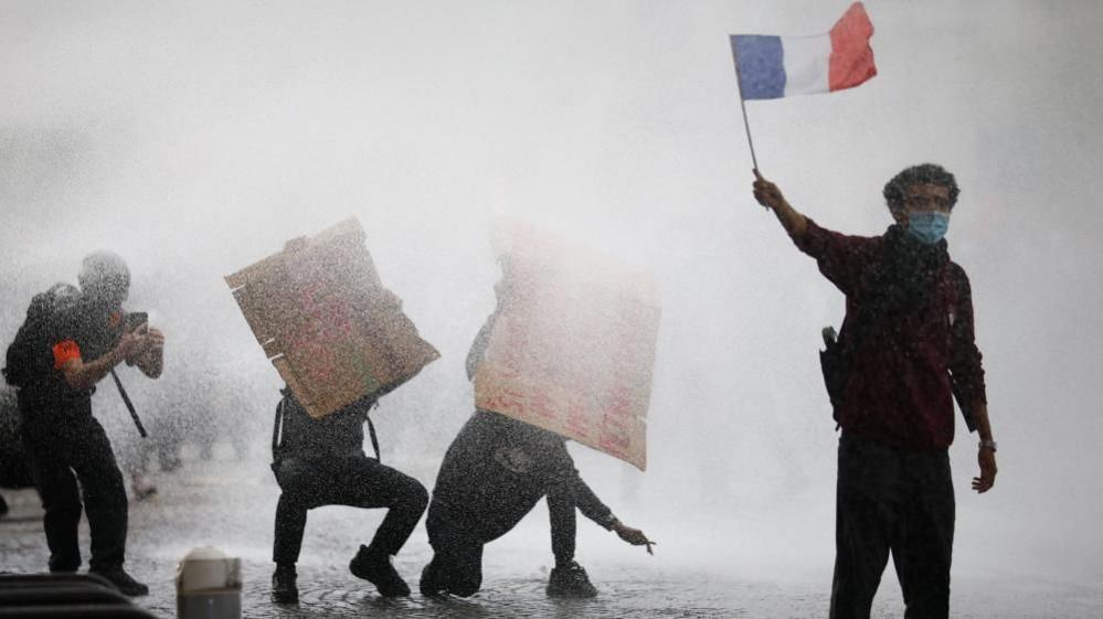 A masked protester holds a French flag as French police use a water canon amid clashes during a demonstration by French high school and university students in Paris.