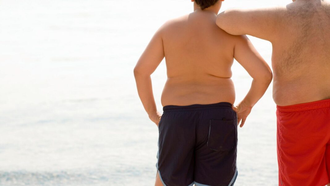 New global guidelines warn against obesity drugs for children under 10 A boy and his father stand in their swimming trunks looking at the sea. Both are overweight or obese
