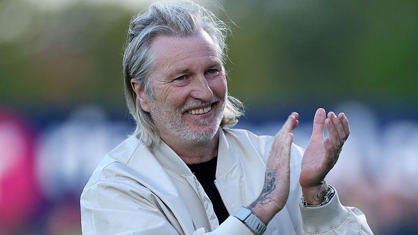 Robbie Savage in a white jacket claps his hands and smiles during a match
