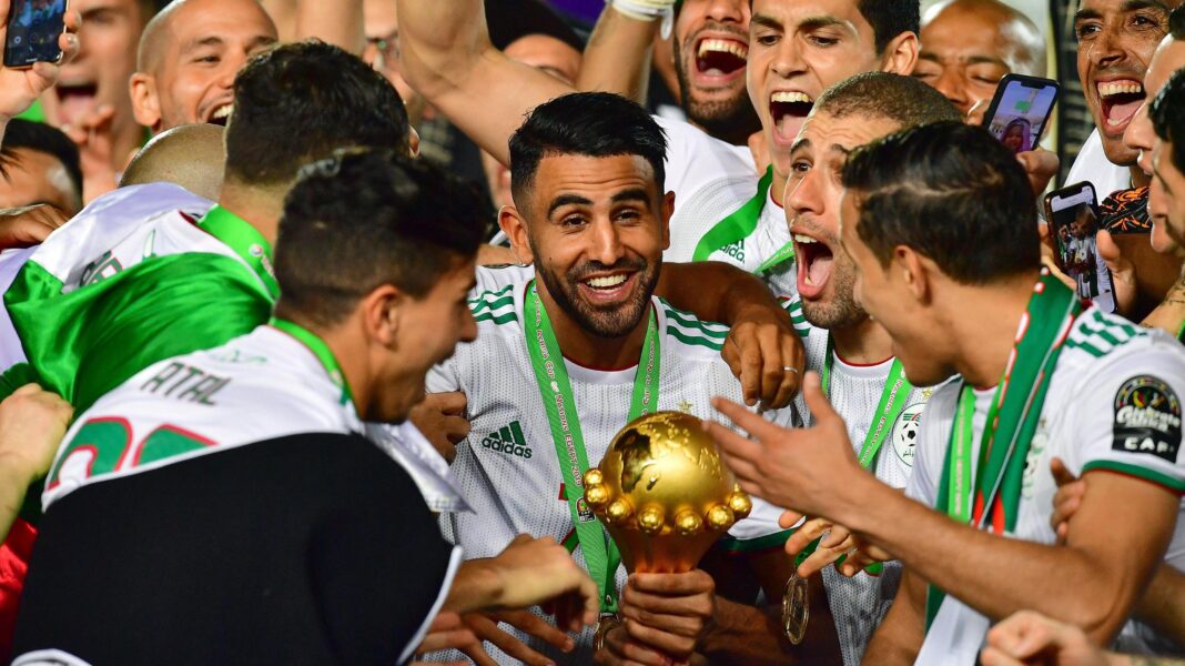 Why has Caf abandoned plans to hold Afcon every four years? Algeria captain Riyad Mahrez, wearing a white shirt and winner's medal on a green ribbon, smiles as he holds the Africa Cup of Nations trophy while surrounded by gleeful team-mates who are celebrating with him after the trophy lift at the 2019 Afcon final