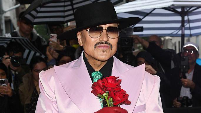 Willy Chavarria on a red carpet with photographers all around wearing a black hat, sunglasses and a pale pink tuxedo with exaggeratedly large lapels and holding three red roses