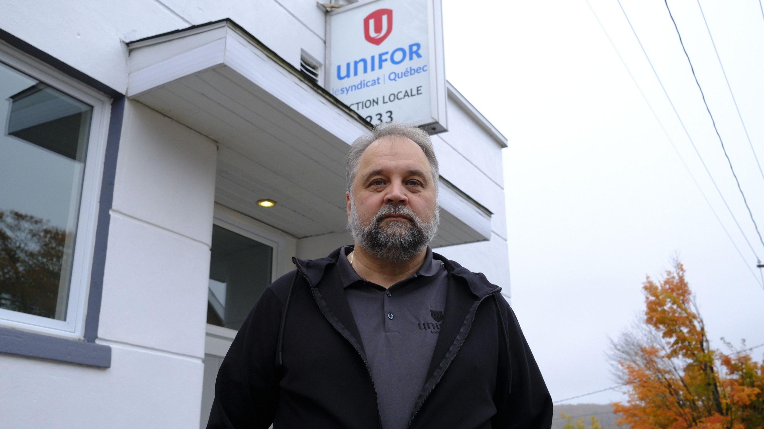 An image showing a man, Stephane Lefebvre, outside a small building with a sign that reads 'UNIFOR Syndicat Quebec'. The man is older with grey hair and a beard, is looking right at the camera and is wearing a black zip up jacket on top of a dark grey polo shirt.