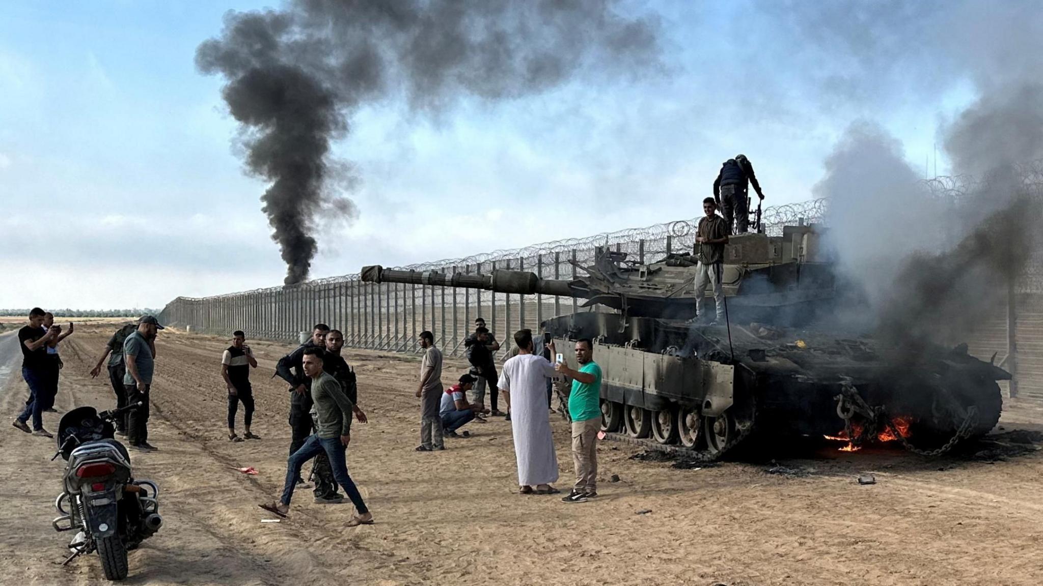 File photo showing Palestinians gathered around an Israeli tank set on fire during the Hamas-led 7 October 2023 attack on Israel, on the Israeli side of the Gaza border fence