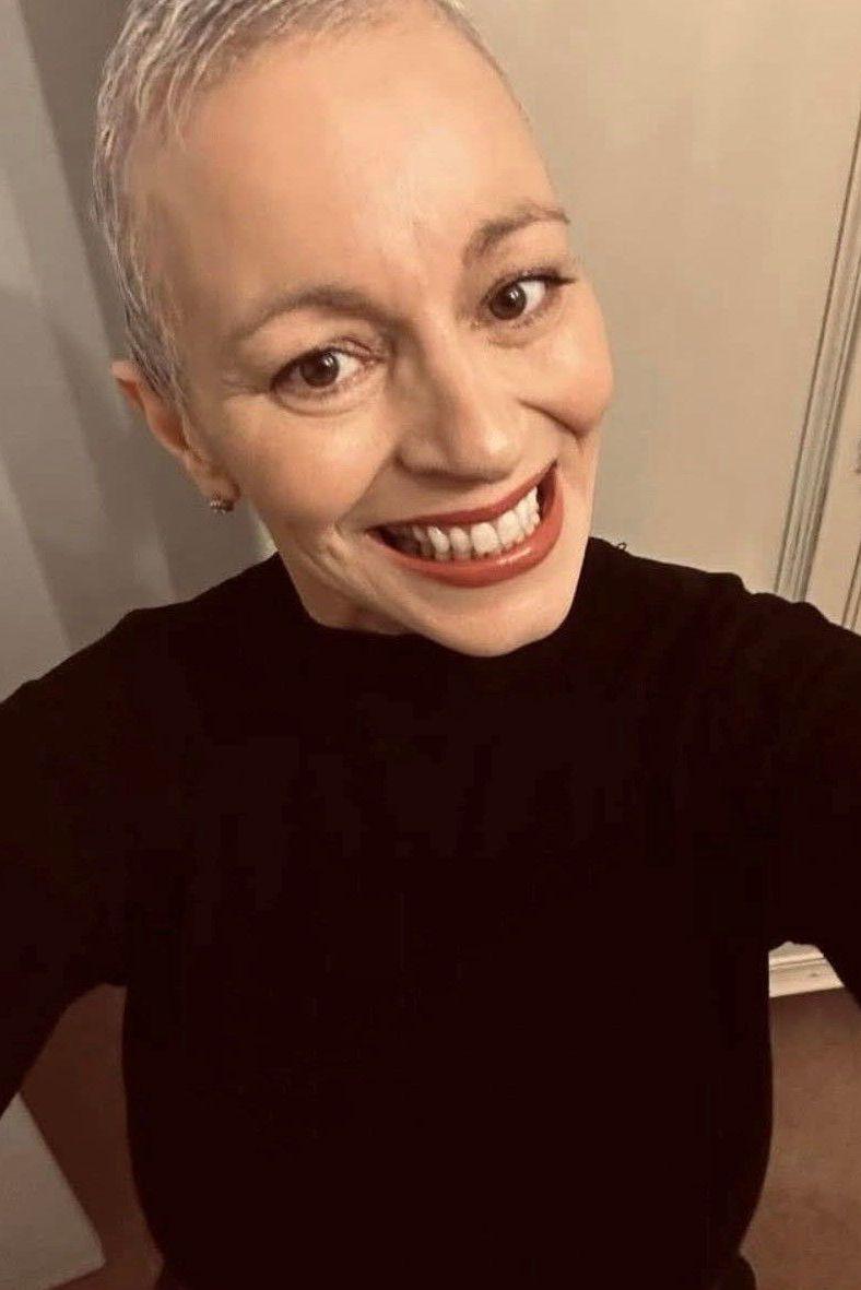 Caroline is seen smiling at the camera in a selfie. She has cropped grey hair, with dark eyes and eyebrows. She is wearing black jumper