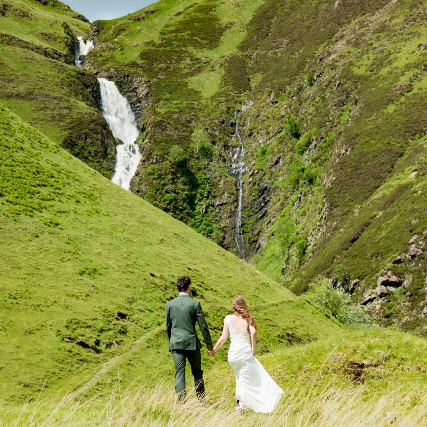 Angelika is in her wedding dress and Patryk is in his green wedding suit as they stand gazing at a waterfall.