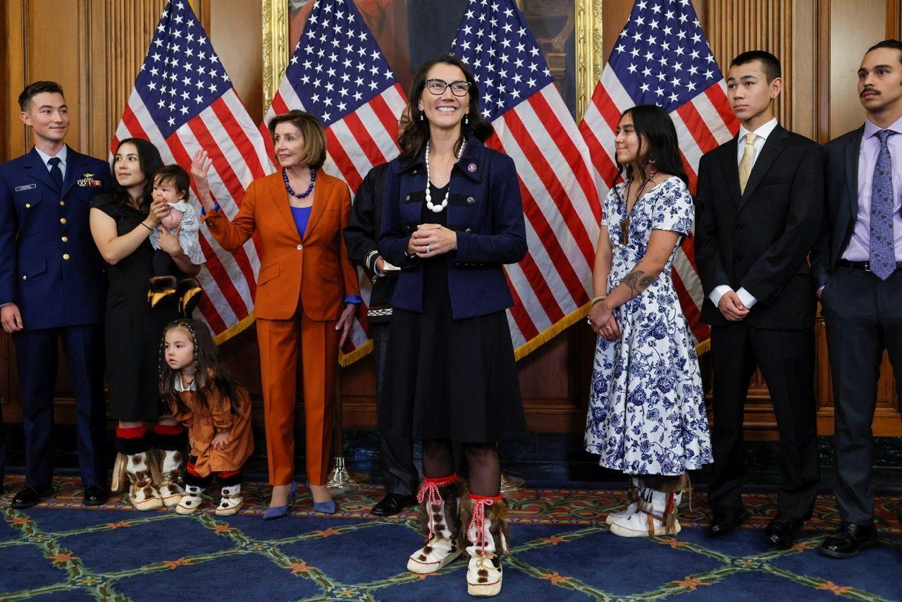 Mary Peltola poses for the cameras after her swearing-in ceremony in front of several US flags. She is wearing traditional boots of the Yup'ik people. Family members and supporters stand around her, including adults and children, some wearing traditional-style footwear and clothing. Nancy Pelosi, in an orange trouser suit, stands nearby.