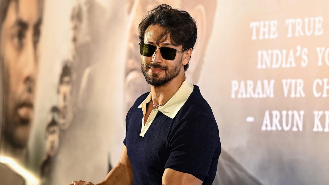 Attack on actor Tiger Shroff is dark reminder of Bollywood’s gangster past Tiger Shroff poses at a promotional event, wearing dark sunglasses and a navy polo shirt with a white collar. The actor leans on a display stand in front of a backdrop featuring large portrait images and text from a film campaign.
