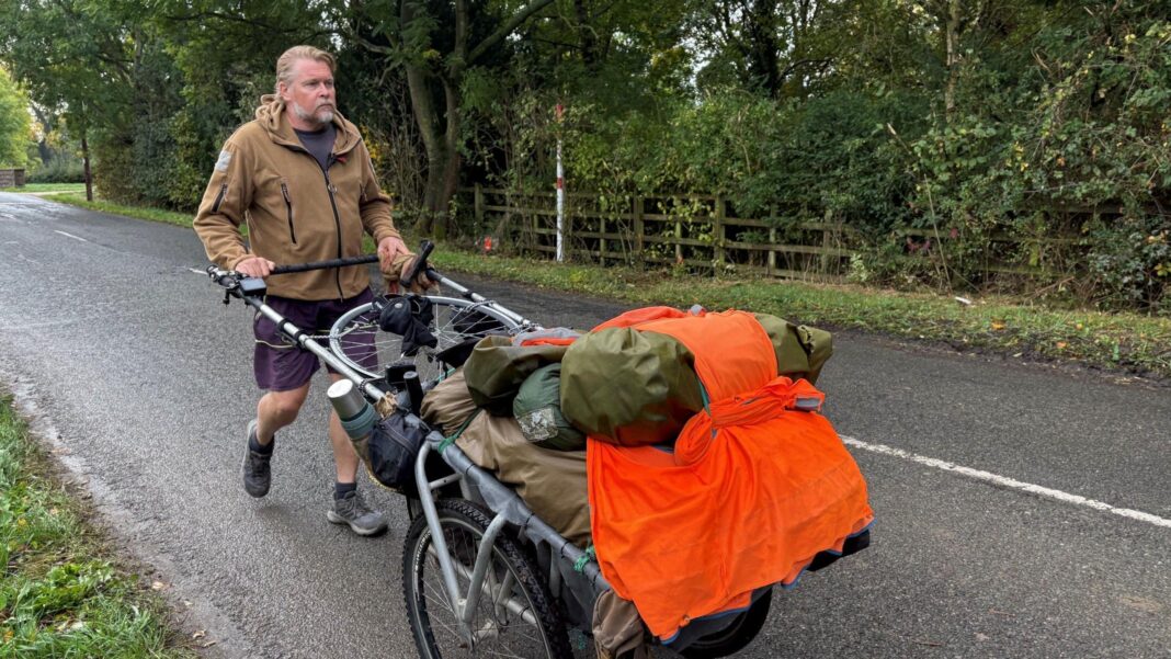 Karl Bushby, wearing a fleece and shorts, pushes a handcart along a fast rural road. An orange hi-vis bib is attached to the front of the cart.