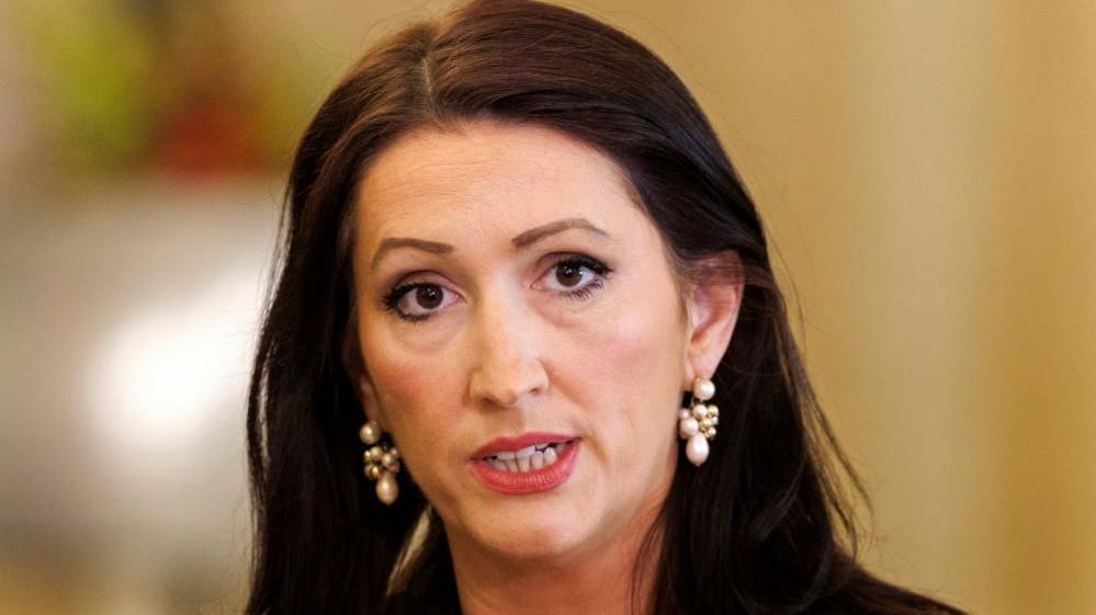 A close-up image of Emma Little Pengelly speaking to reporters. She has dark hair and is wearing white pearl earrings.