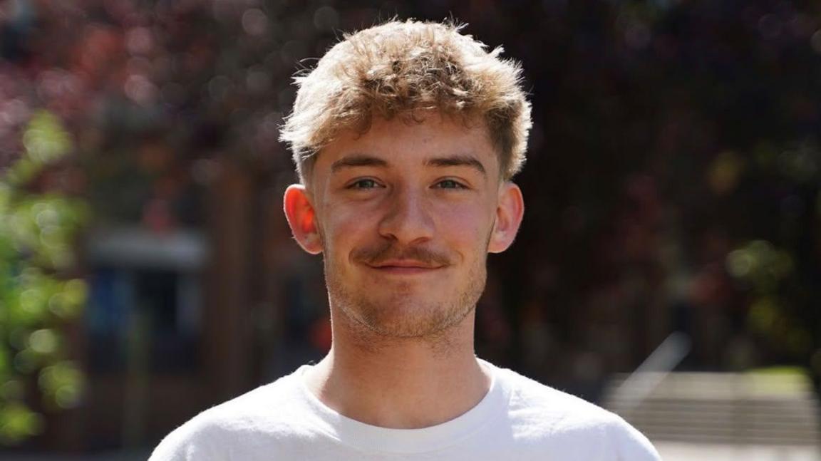 Head and shoulders shot of Lewis Wilson, a young man in a white T-shirt, with a moustache and light beard