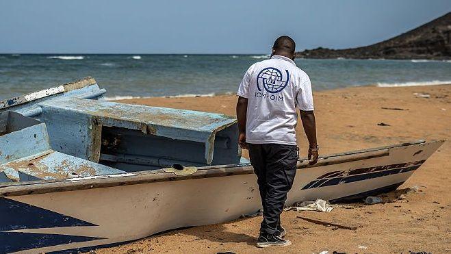 boat wreckage on the beach. A man wearing a white IOM tshirt, is standing to its right and is looking at it.