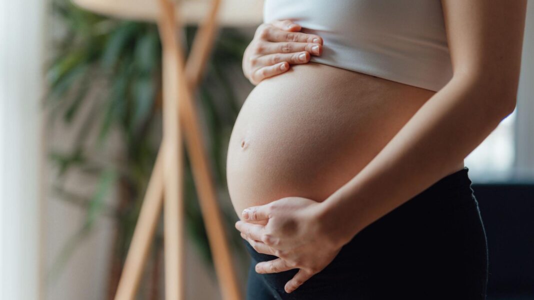 NHS bosses say smaller maternity units may have to close A pregnant woman places her hand on the top and bottom of her baby bump. She is wearing a white cropped top and her pregnant belly is centre of the frame. She is wearing black leggings and is in a living room.
