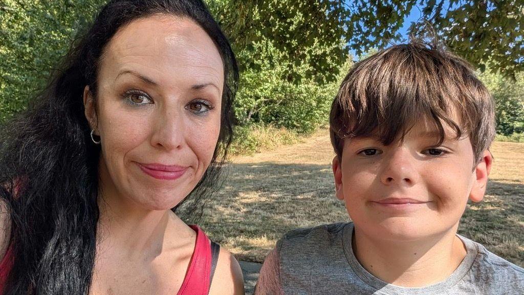 Samantha sits next to her son Oscar. Samantha wears a bright pink vest top and has long dark hair, while Oscar wears a grey T-shirt. Both are facing the camera. Sunlight filters through the leaves overhead, creating patches of light and shadow across the scene. Behind them is a dry, grassy field with green trees and shrubs in the distance under a blue summer sky.