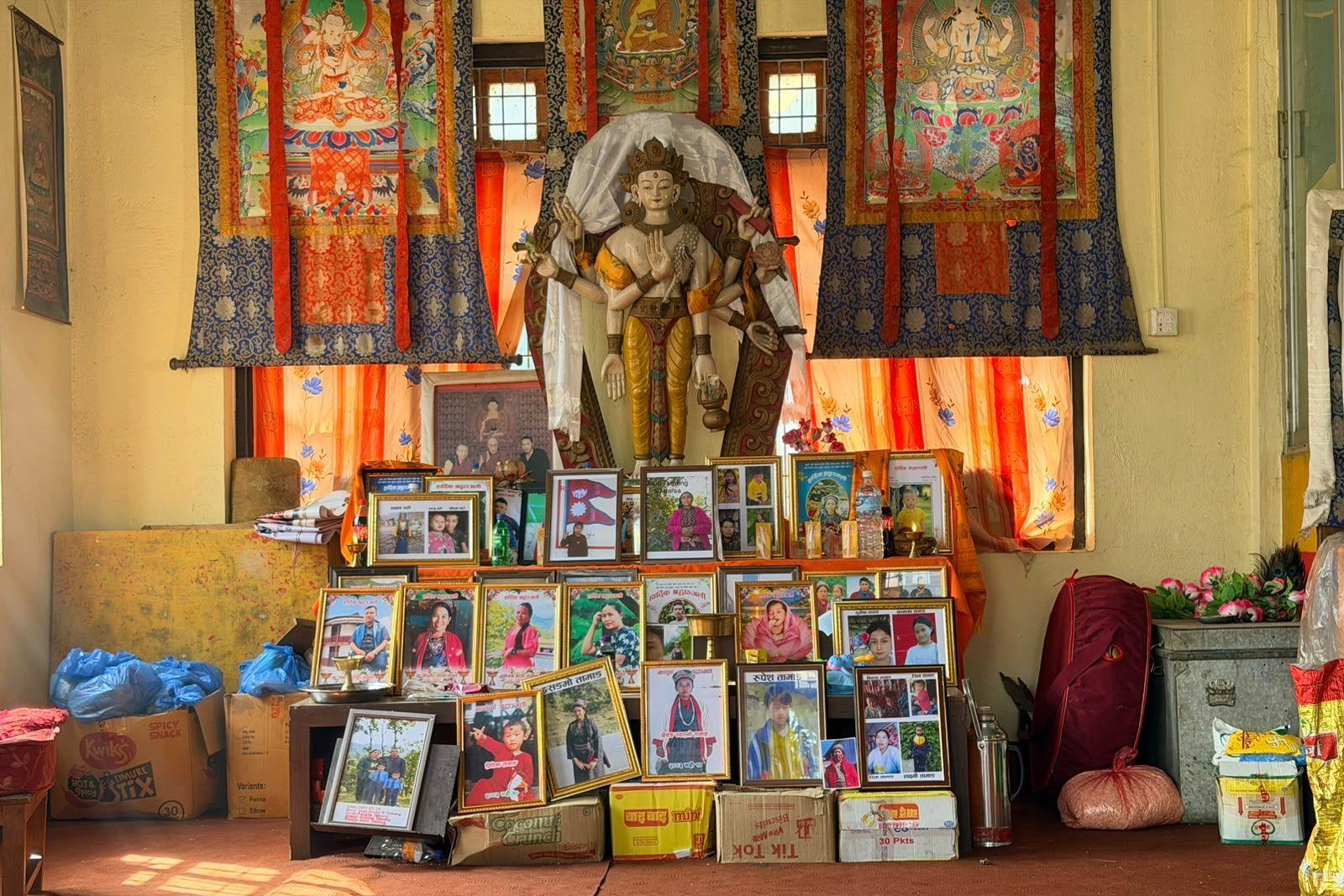 Dozens of framed photographs arranged beneath the figure of a religious deity inside a building, with multicoloured and patterned curtains hanging in the background