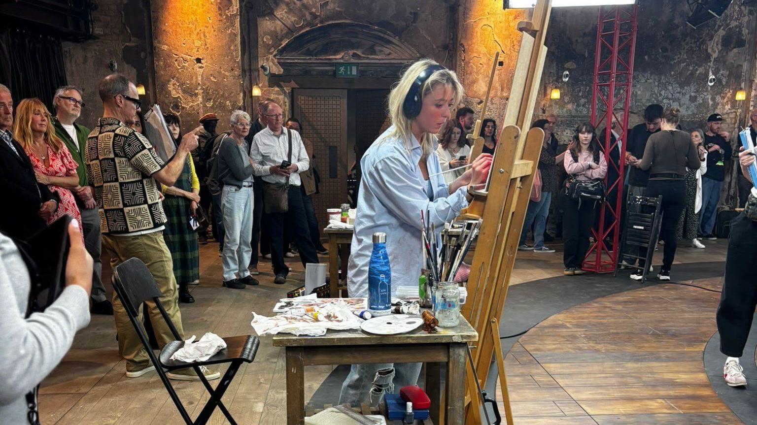 Makenzy Beard paints a celebrity sitter during her time on the Portrait Artist of the Year show. She stands with an easel surrounded by TV crews.
