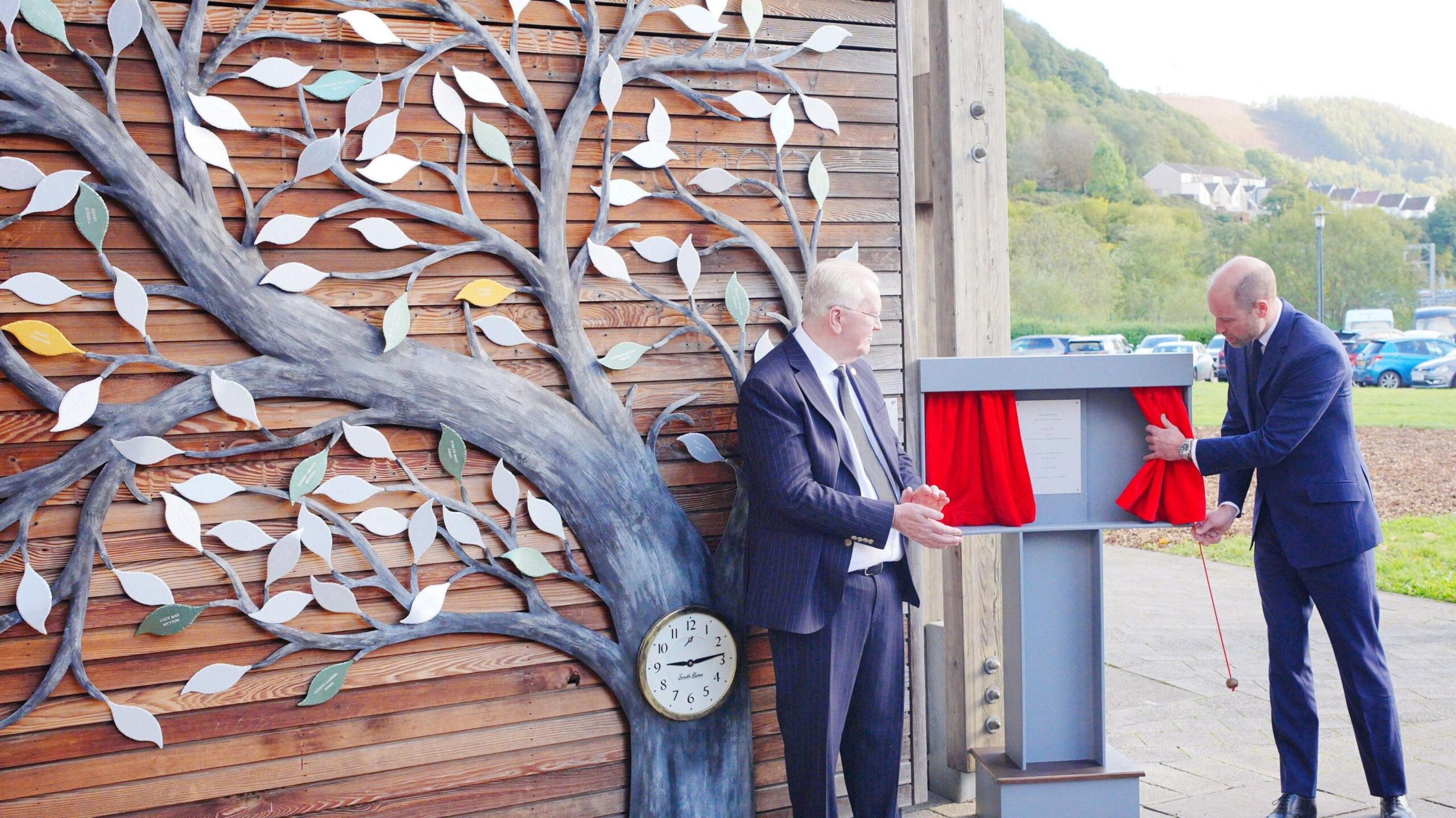 Prince William is pulling back a red curtain which reveals a plaque. Beside him is a large tree sculpture.