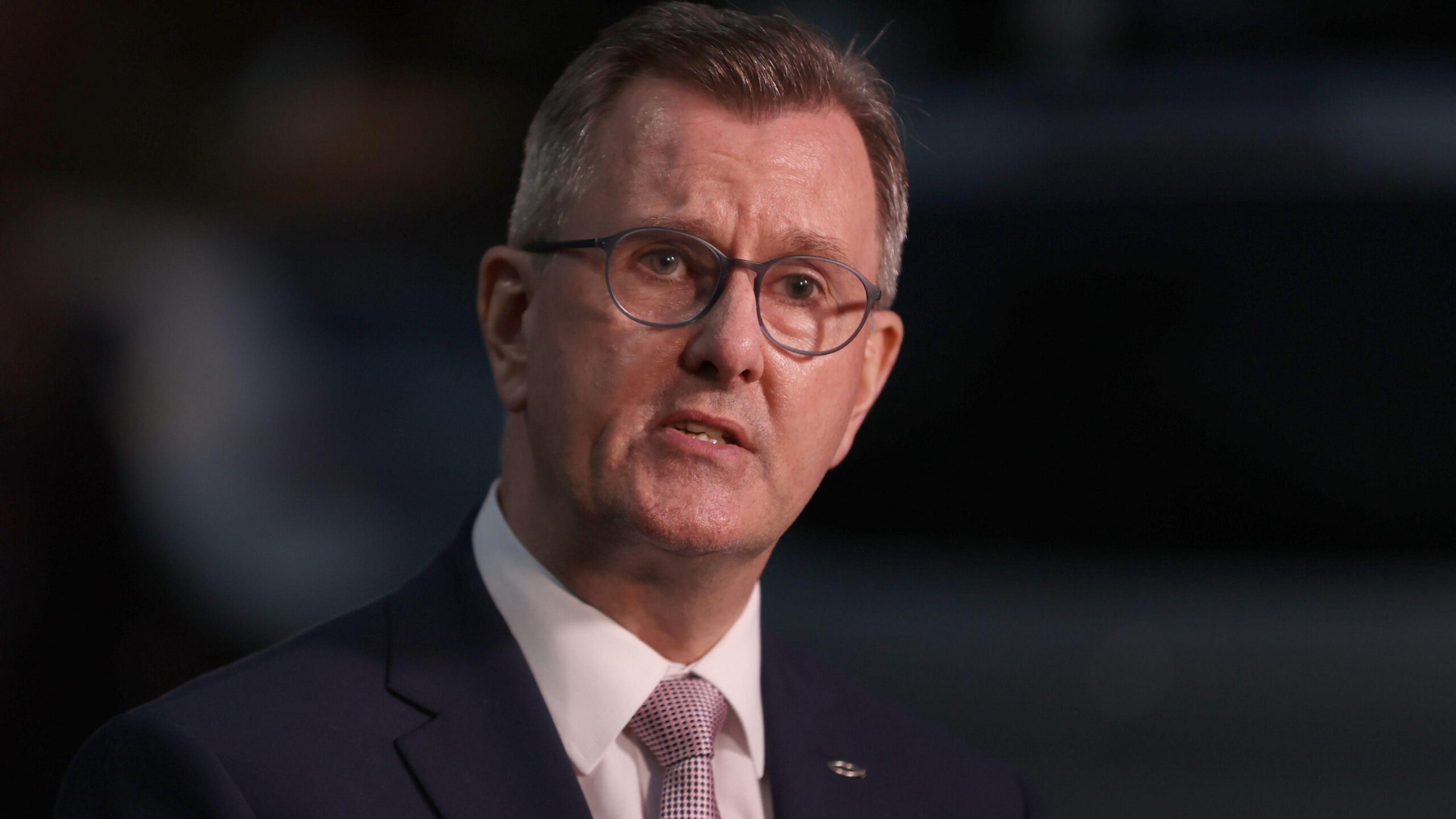 A man is looking sideways, directly at the camera. He has short dark hair and is wearing rounded glasses. The man is dressed in a dark suit jacket with white shirt and light pink tie, which has a textured pattern. There is a small silver fish symbol attached to the lapel of his jacket.