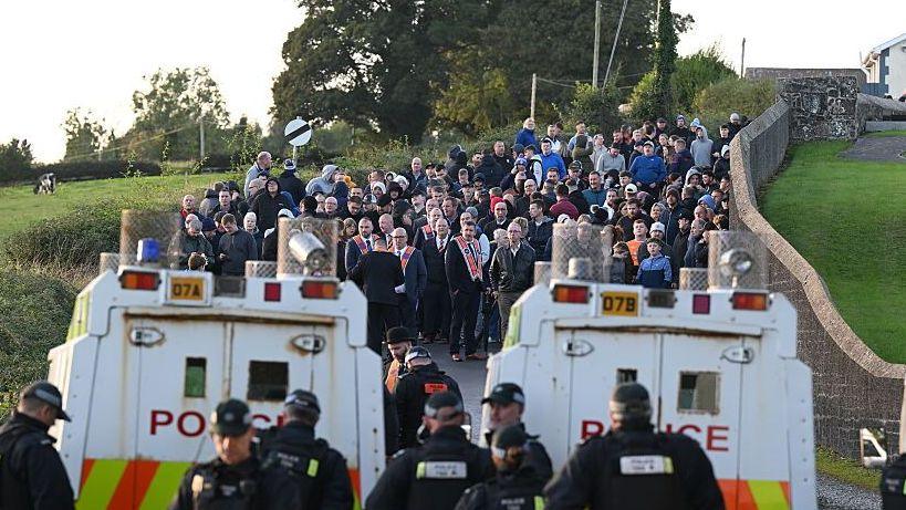 Two police Land Rovers parked in front of a crowd of people gathered on a country road.