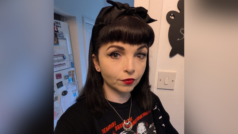 A woman with black hair and a fringe wears a black t-shirt, red lipstick and has a black bow in her hair in a photo taken in a kitchen in front of a fridge.