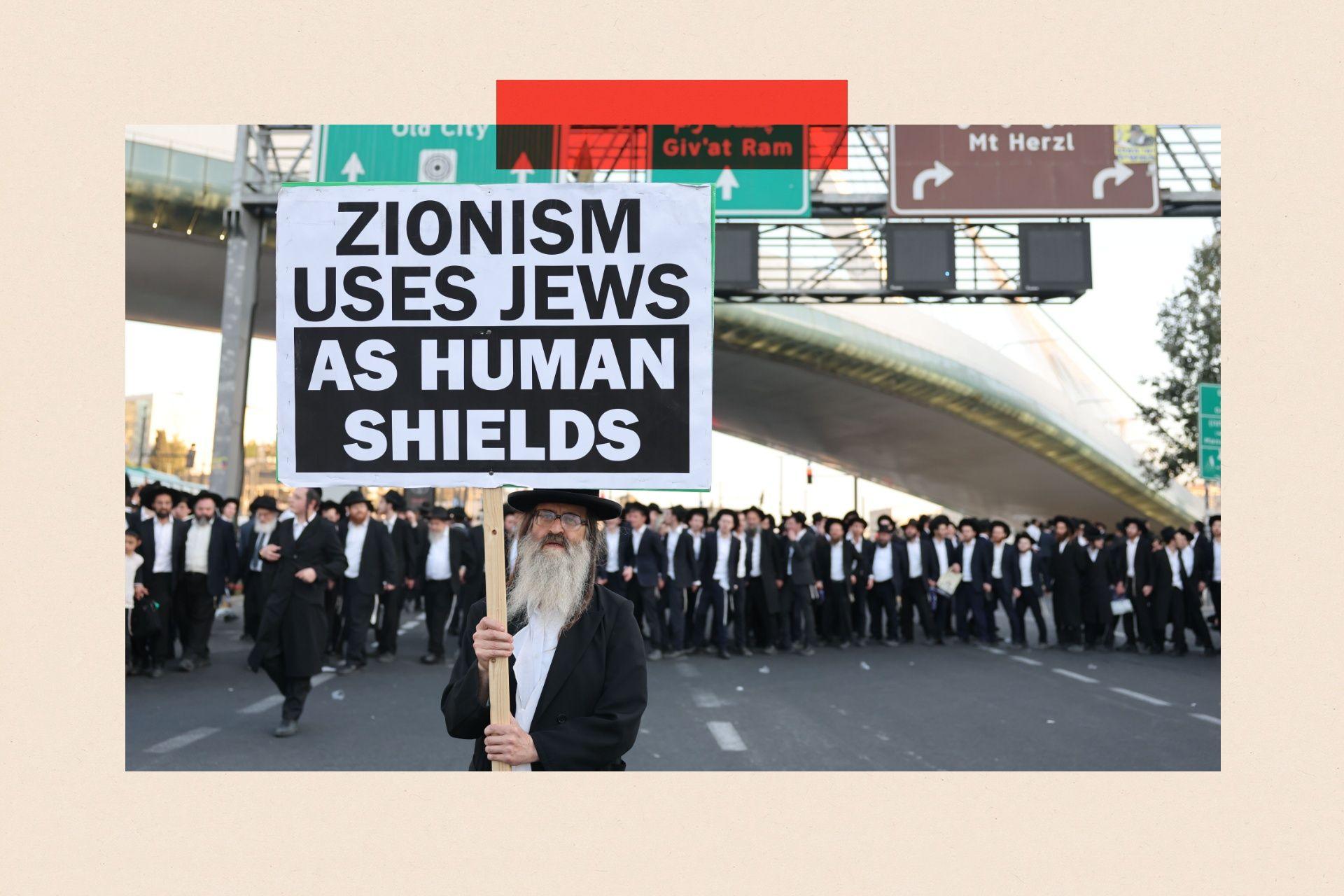 A Haredi man hold a sign saying 'Zionism uses Jews as human shields' under a motorway overpass, with other Haredi men in the background