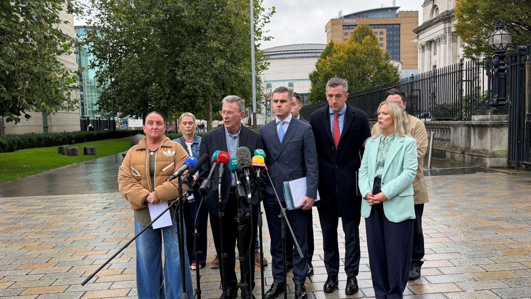A group of eight people stand on light grey and brown bricks outside, in front of a large white stone building.