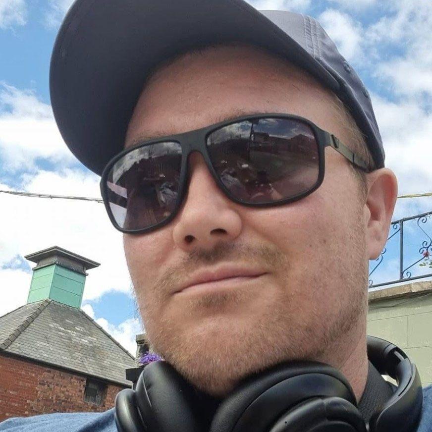 A selfie of a man with a fair beard, wearing a black baseball cap, sunglasses and black headphones around his neck. He has a slight smile as he looks at the camera.
