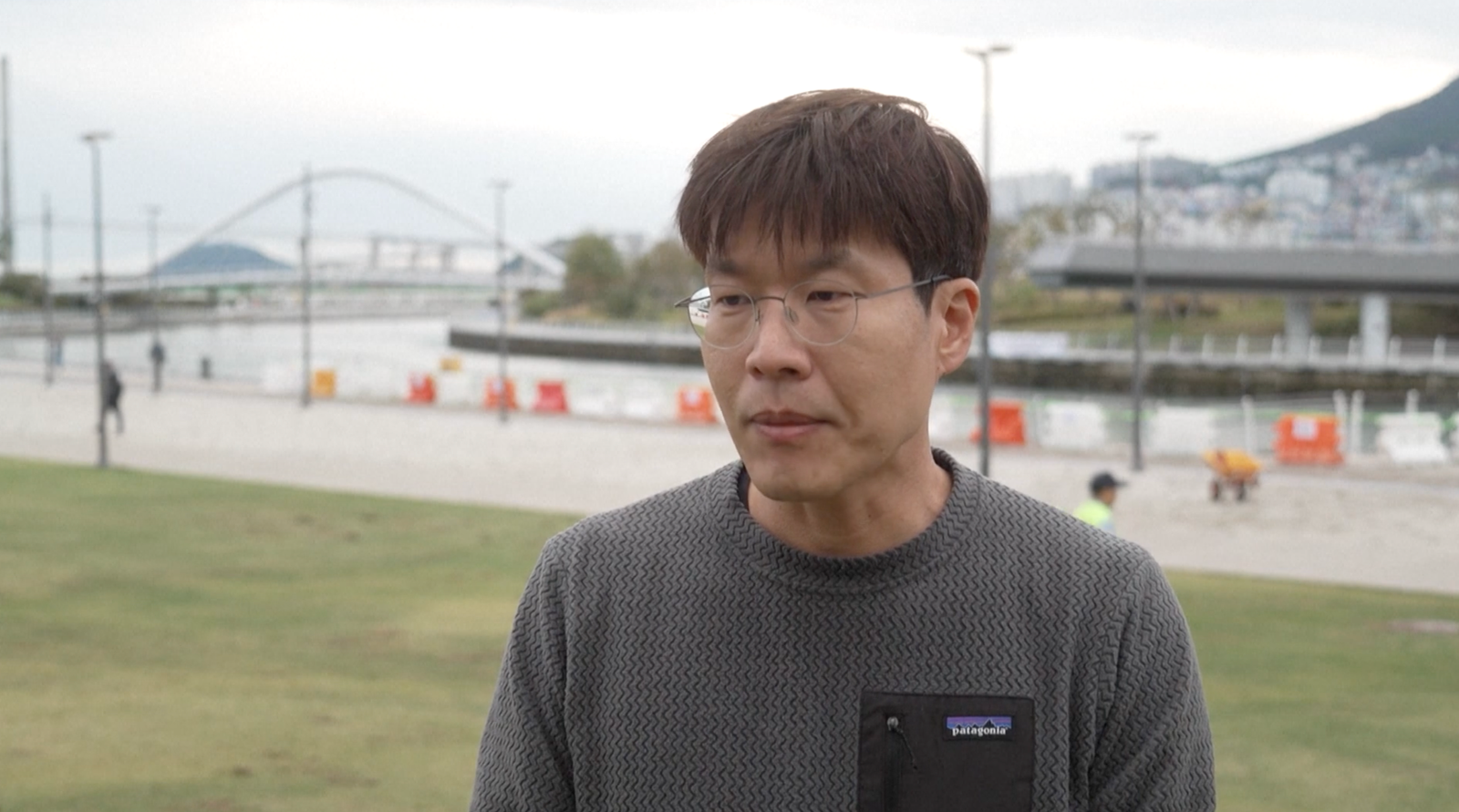 A person wearing glasses and a grey patterned Patagonia sweatshirt stands on a grassy area beside a waterfront canal in Busan, South Korea. In the background, a pedestrian bridge arches over the water, while construction barriers, lampposts and a riverside walkway line the canal. A hill with buildings is visible in the distance under an overcast sky. Visible text on the clothing reads “Patagonia.”
