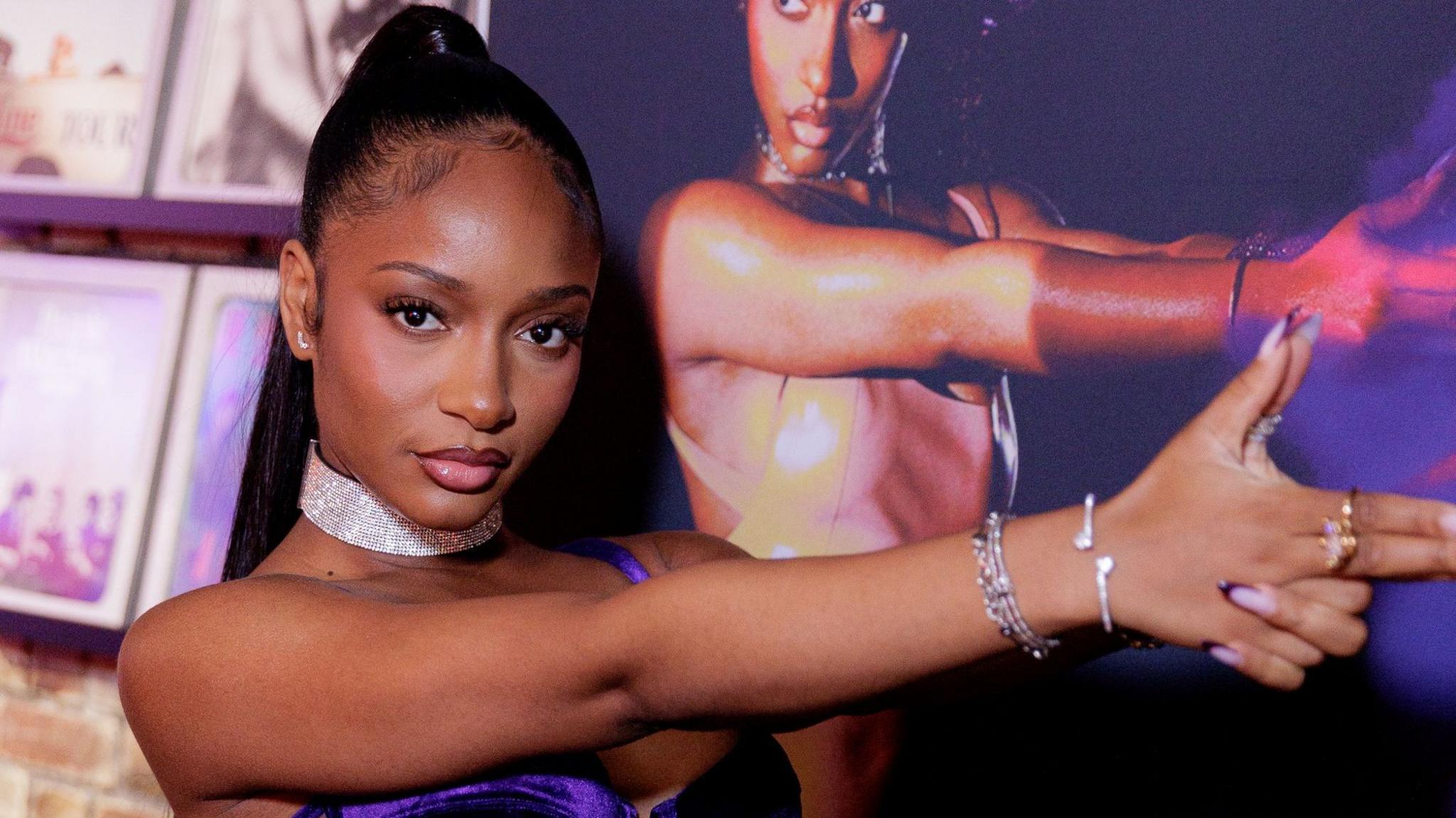Ayra Starr poses with her hands in a gun pointing position. She mimics a poster of her album cover she is behind her.